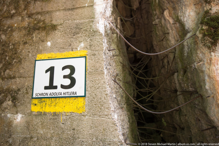 Marker 13 labeled Schron Adolfa Hitlera beside a jagged concrete fracture crossed by exposed steel reinforcement.