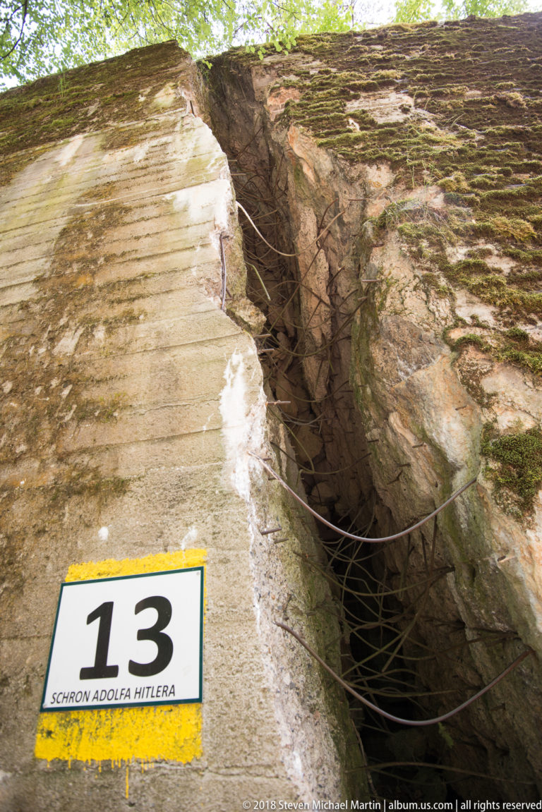 Close view of deep crack between concrete bunker masses, exposed reinforcing bars, and marker 13 reading Schron Adolfa Hitlera.