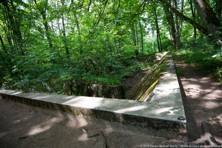 Long rectangular low concrete foundation enclosing a sunken space beside a forest path.