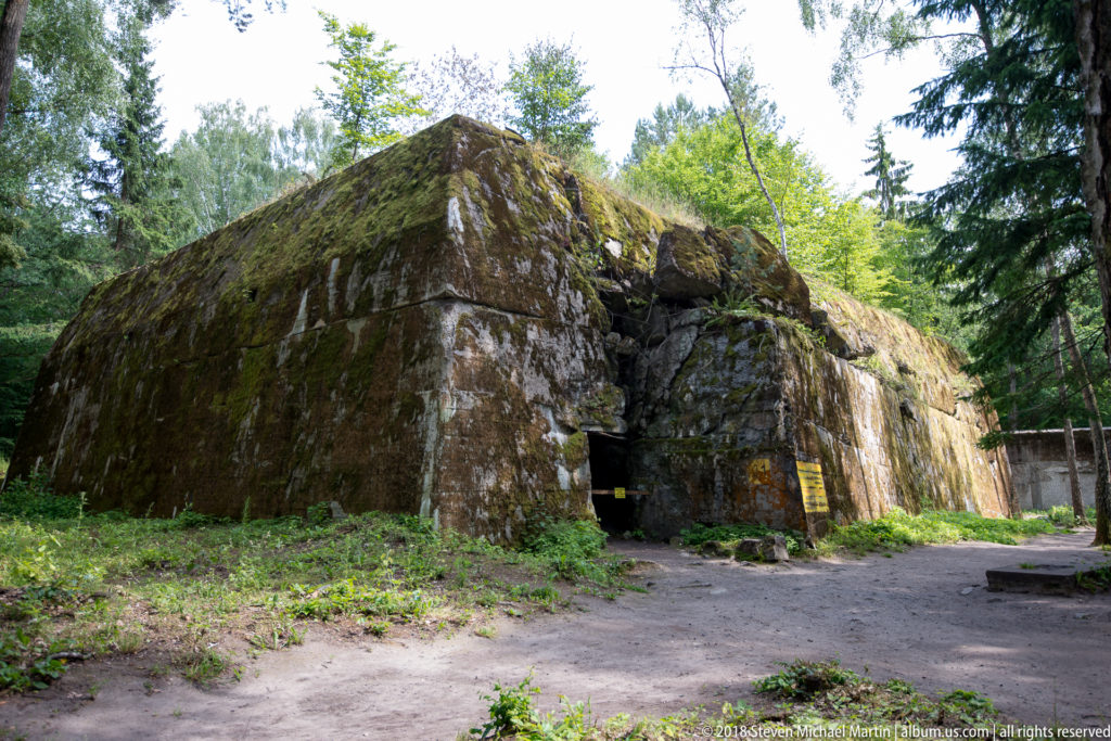 Low moss-covered concrete bunker with thick sloping walls and a recessed entrance in a forest clearing.