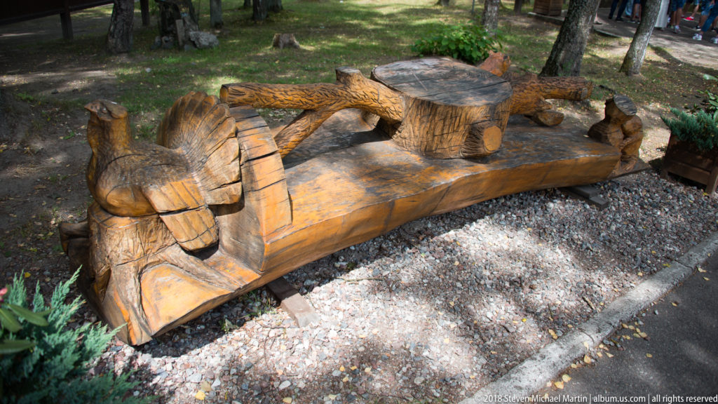 Heavy carved wooden bench with animal-shaped supports and woodland motifs standing on gravel among trees.
