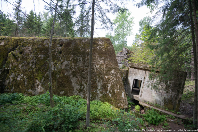 Moss-covered concrete bunker ruins with a massive wall standing beside a tilted section and dark opening in forest.