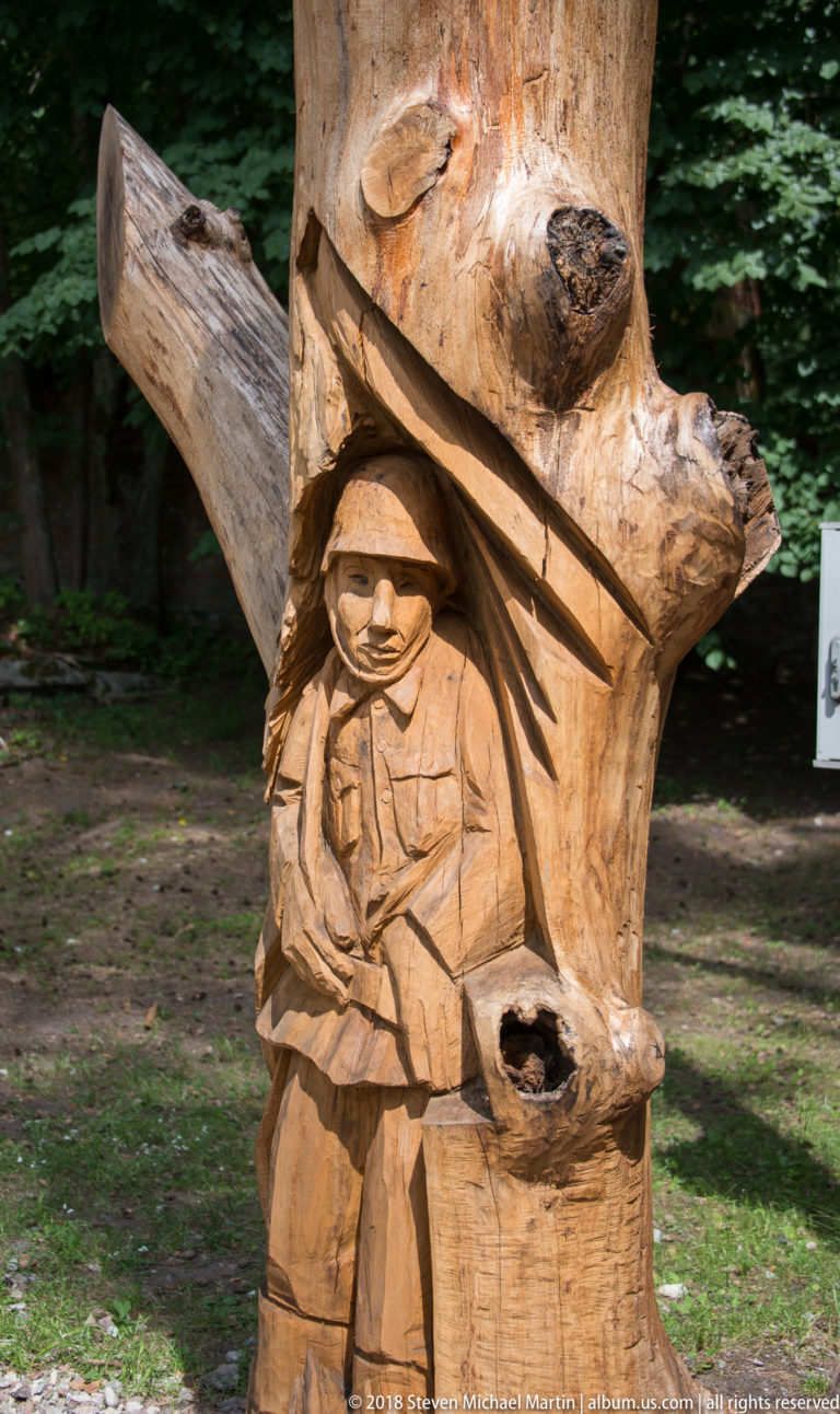 Small wooden soldier figure carved into a tree trunk beneath a projecting branch-like form.