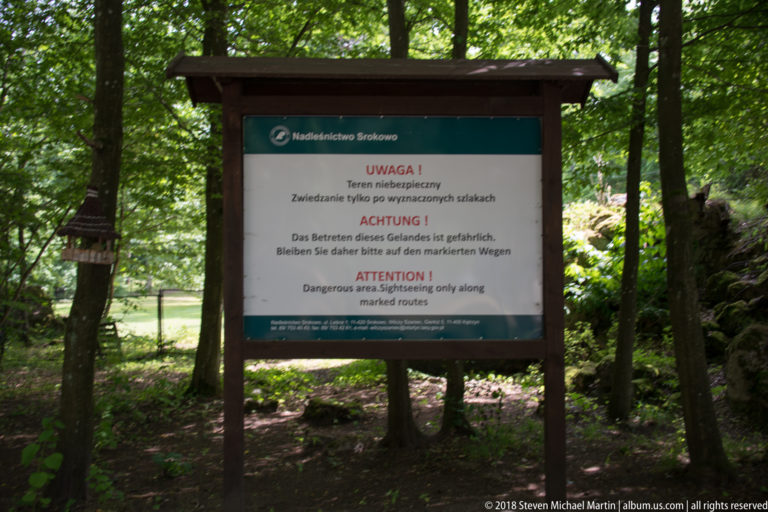 Large woodland warning sign in Polish, German, and English telling visitors that terrain is dangerous and to stay on marked routes.