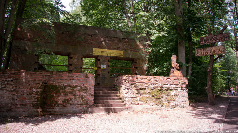 Brick ruin with steps and rectangular openings, interpretive sign above, and wooden directional signs among trees.