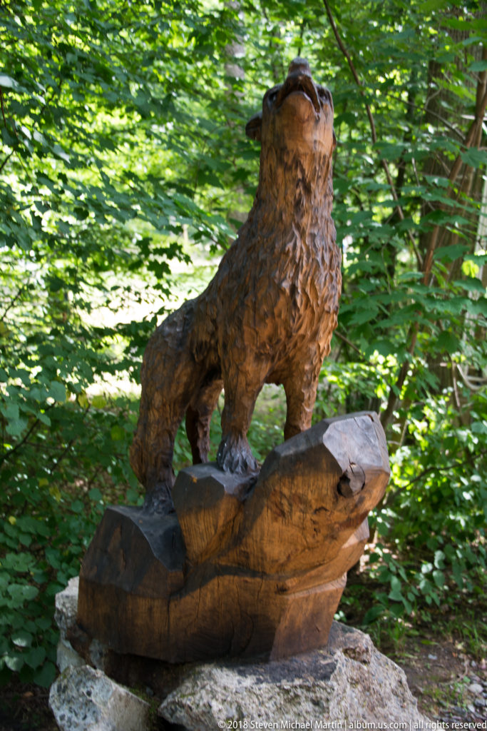 Carved wooden wolf with raised head standing on a sculpted log base against green woodland.