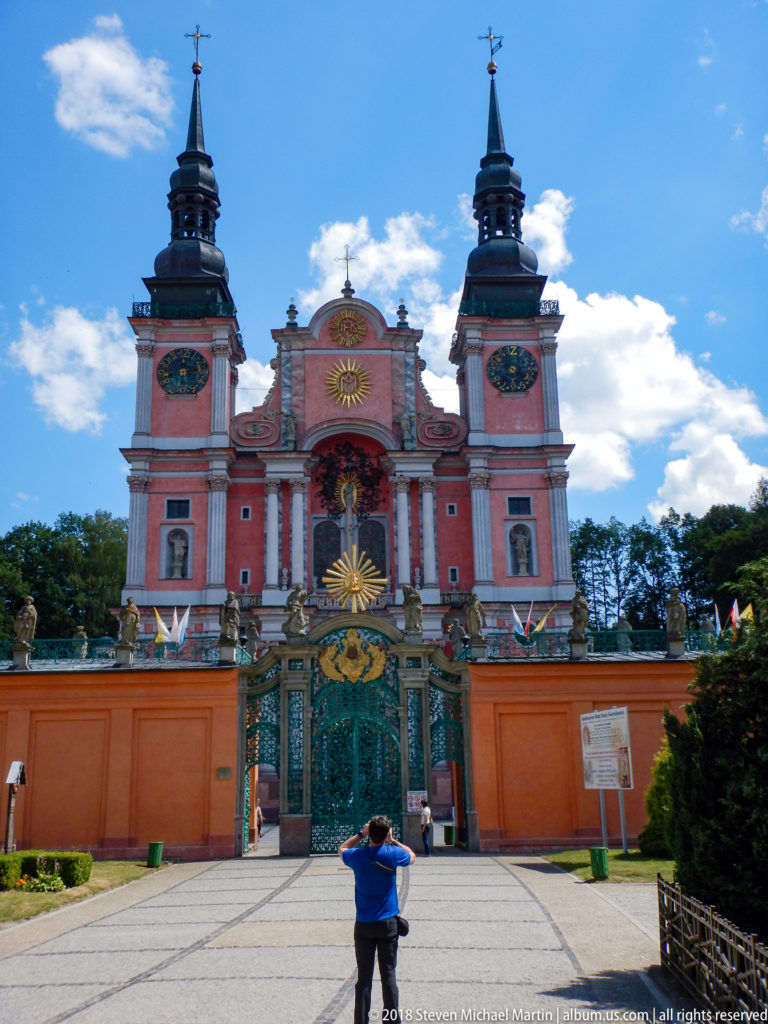 Visitor photographing the pink twin-towered sanctuary from the paved approach, facing its ornate green and gold entrance gate.