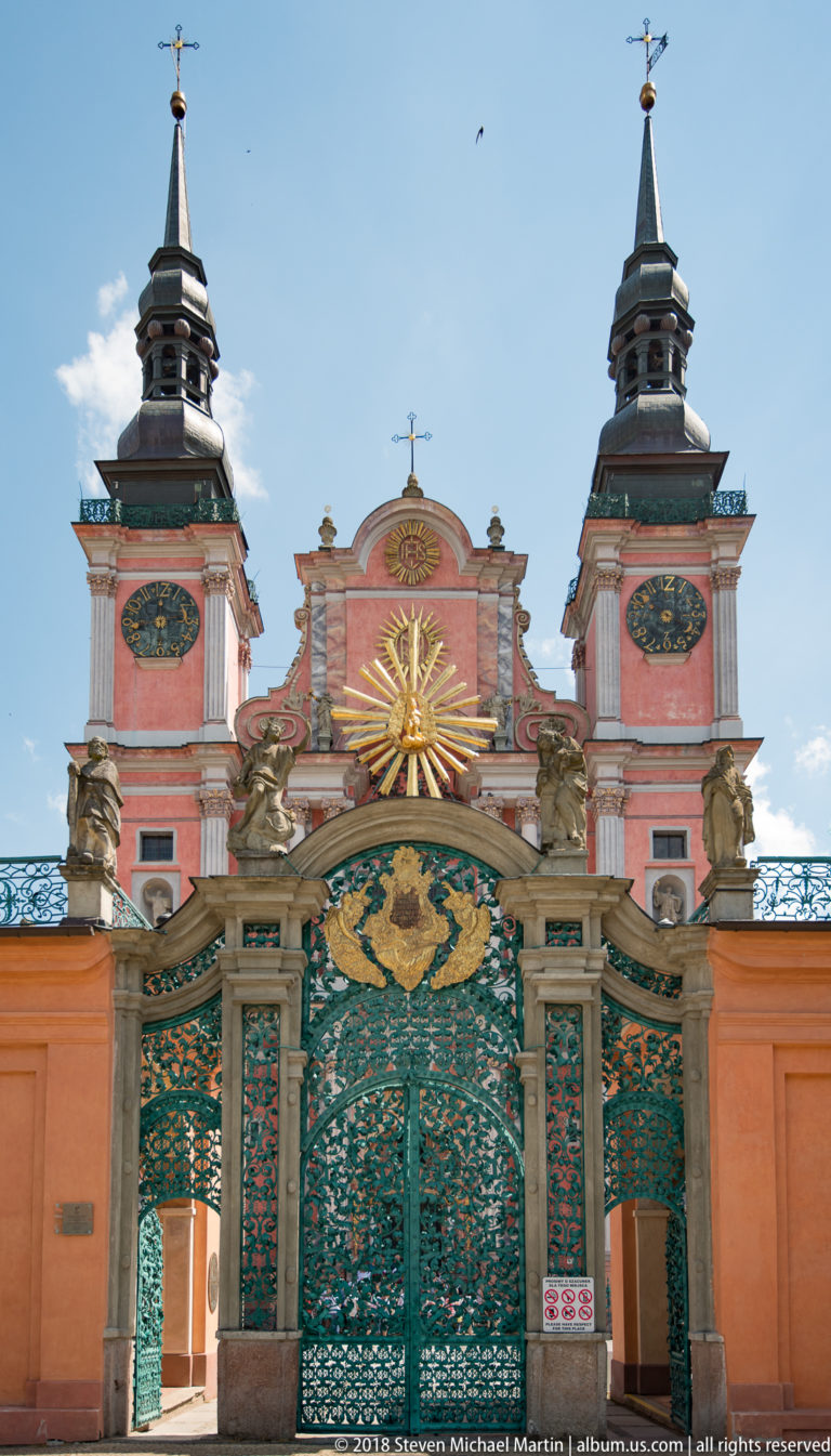 Ornate green and gold entrance gate beneath the pink twin-towered church façade, with sculptures and a central gilded sunburst.