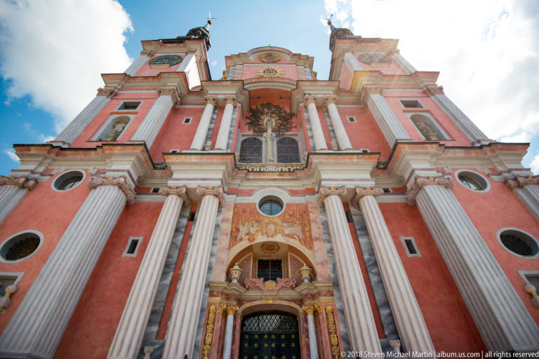 Steep upward view of the pink Baroque church façade with white columns, ornate portal, upper window, and curved gable.