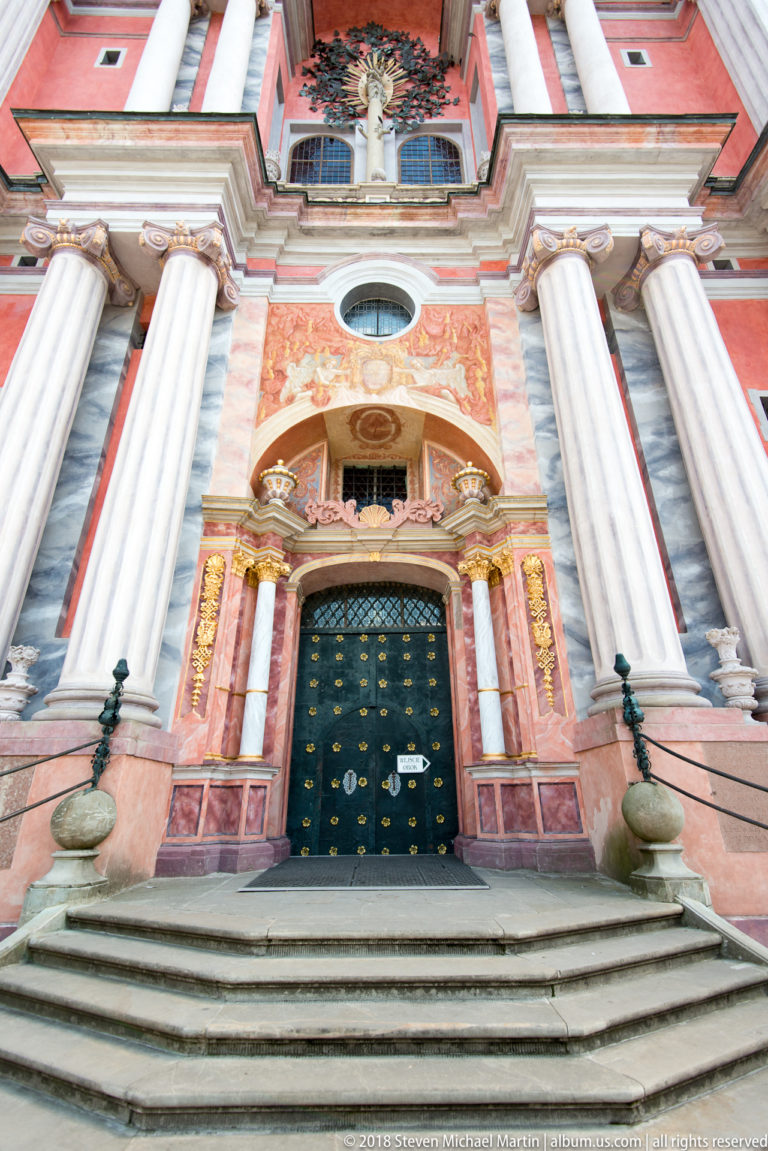 Frontal low-angle view of broad steps leading to a dark church door framed by tall columns and gilded Baroque ornament.