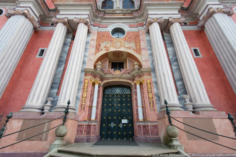 Low-angle view of the sanctuary entrance with tall paired columns, dark studded doors, gilded ornament, and curved stone steps.