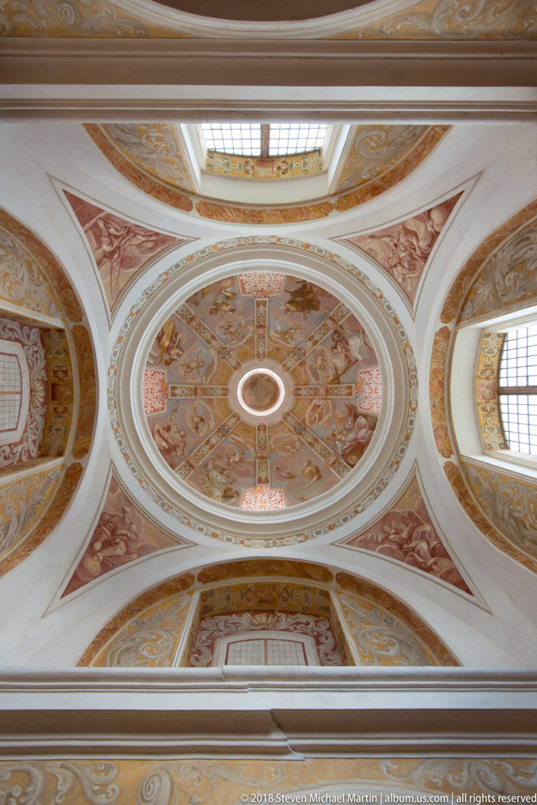 Upward view of a frescoed church vault with oval compartments, pale ribs, gilded ornament, and painted religious scenes.