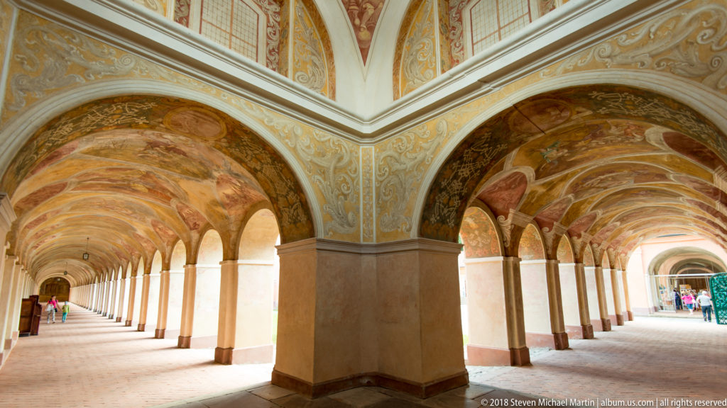 Corner of a frescoed cloister with a large square pier and two intersecting arcaded walkways opening toward daylight.