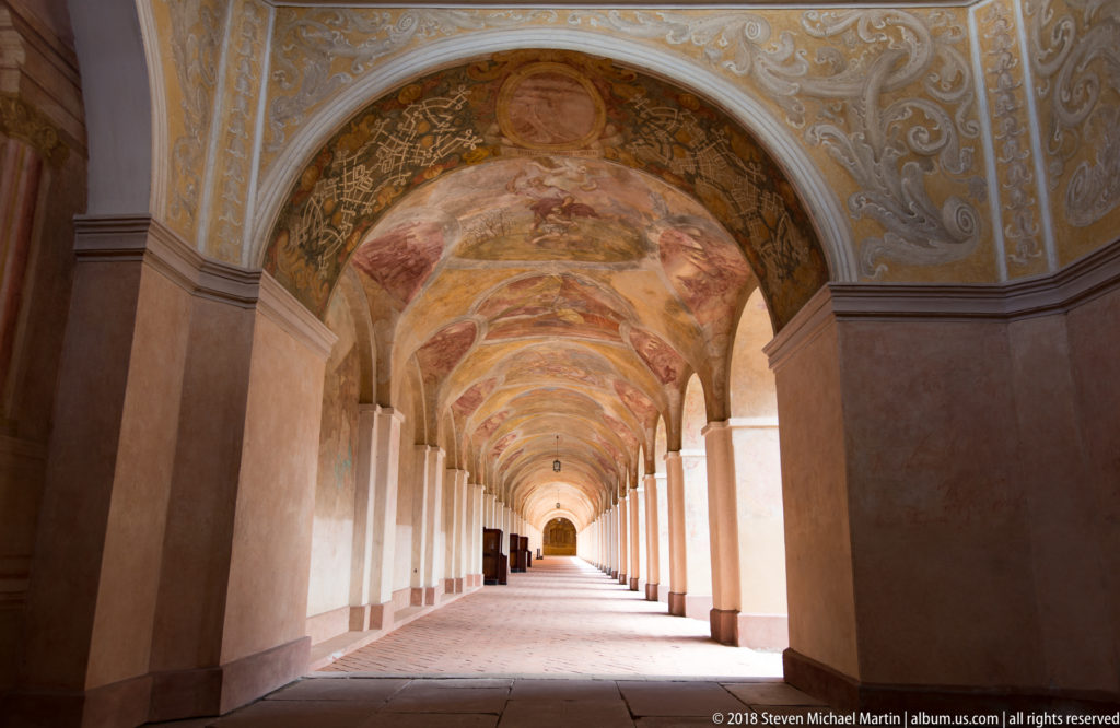 Long covered cloister corridor with repeated arches, pink piers, frescoed vaults, pale walls, and a bright opening at the far end.