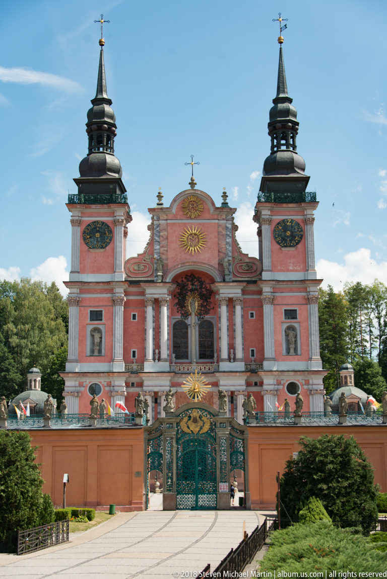 Full frontal view of the red Baroque sanctuary with twin towers behind an ornate gate and central approach path.