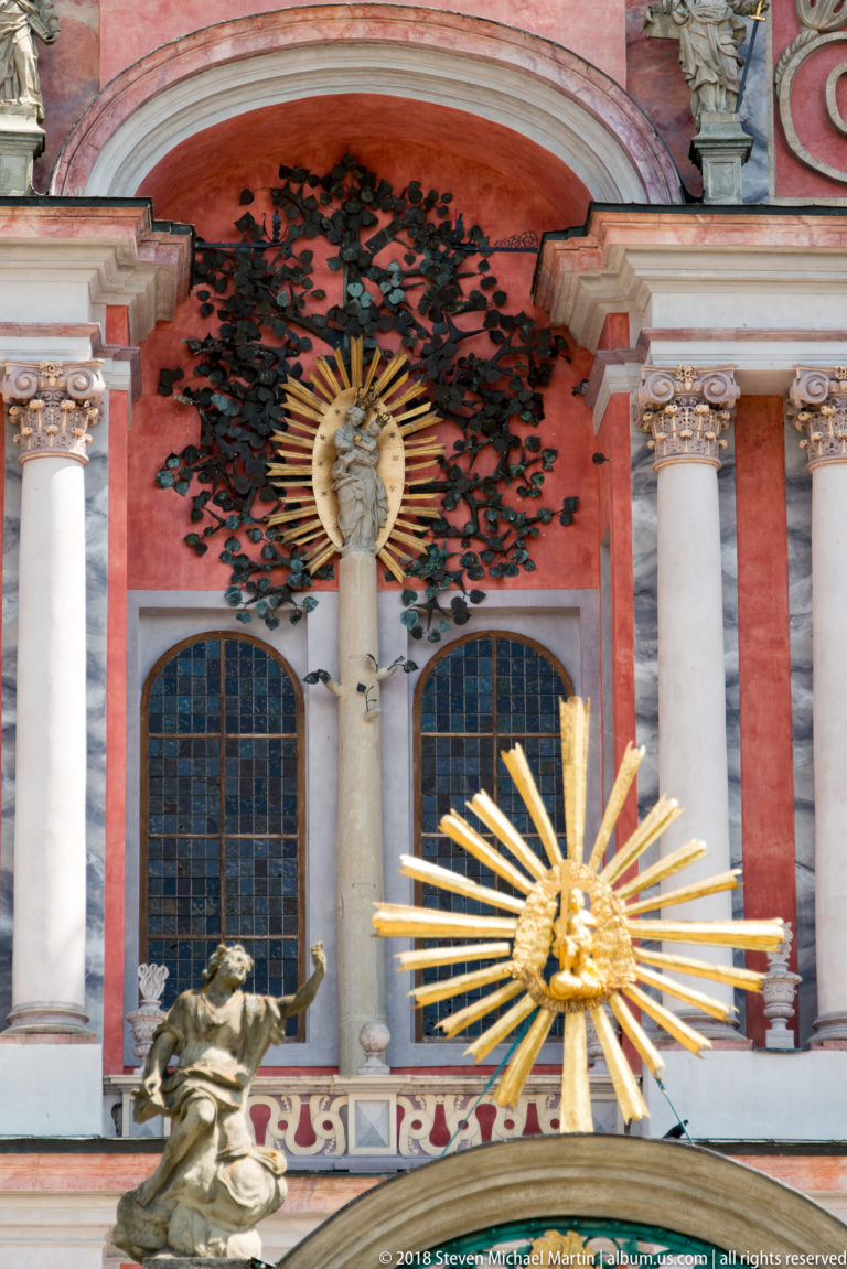 Central red façade detail with dark foliage, pale Marian relief, white columns, sculpture, and a large gilded sunburst in front.