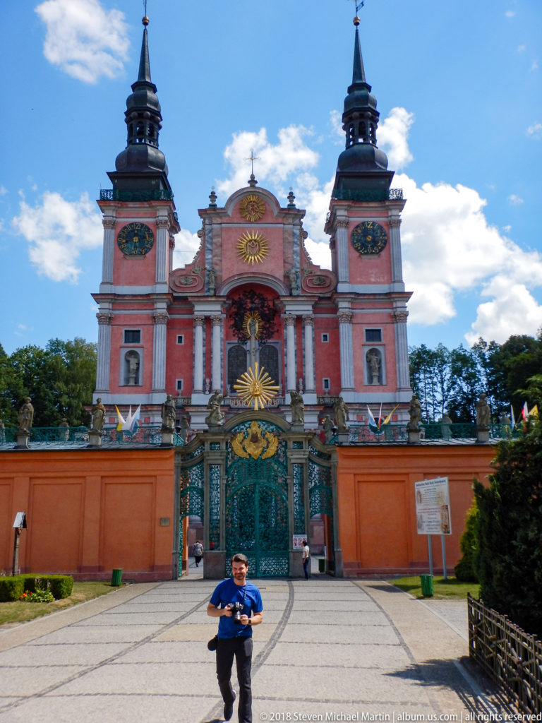 Frontal view of the twin-towered pink sanctuary and ornate gate, with a visitor walking across the paved forecourt.