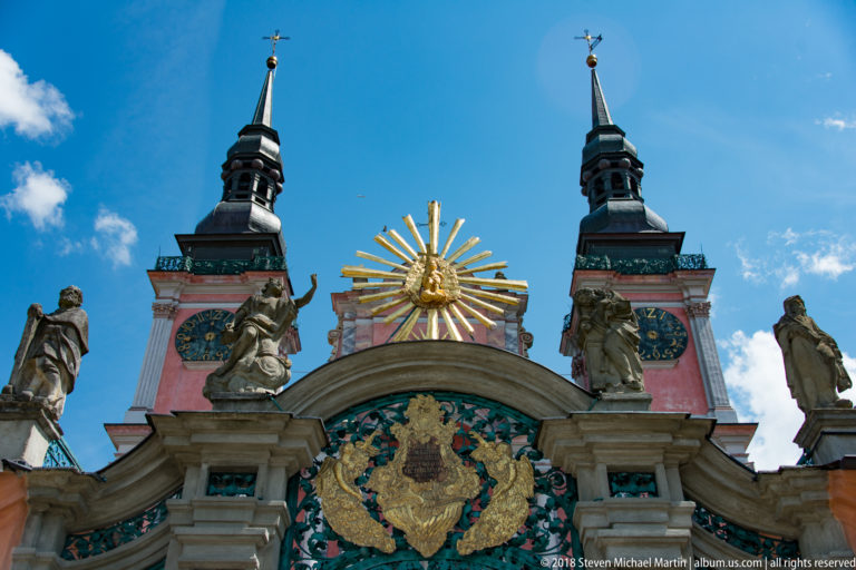 Low-angle view of the red-and-white sanctuary façade with twin towers, clocks, statues, gilded rays, and central emblem.