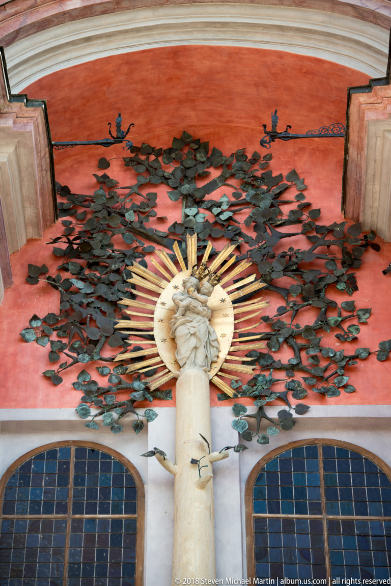 Exterior red façade detail with dark metal foliage surrounding a pale Marian relief and gilded sunburst above two arched windows.