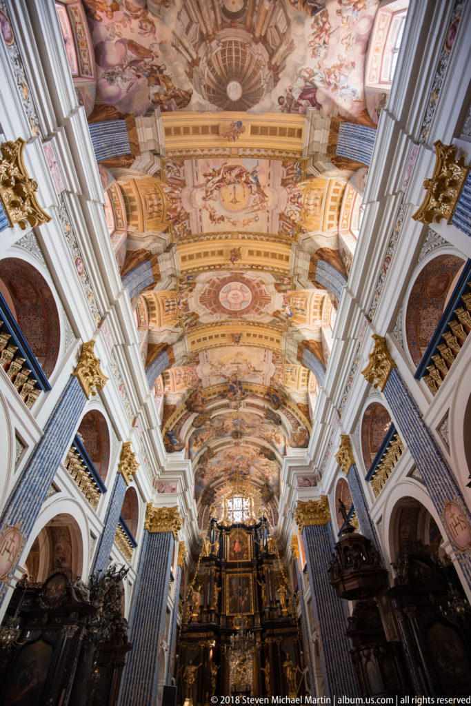 Axial nave view dominated by frescoed vaults, with blue patterned pilasters, gilded capitals, dark furnishings, and distant altar.