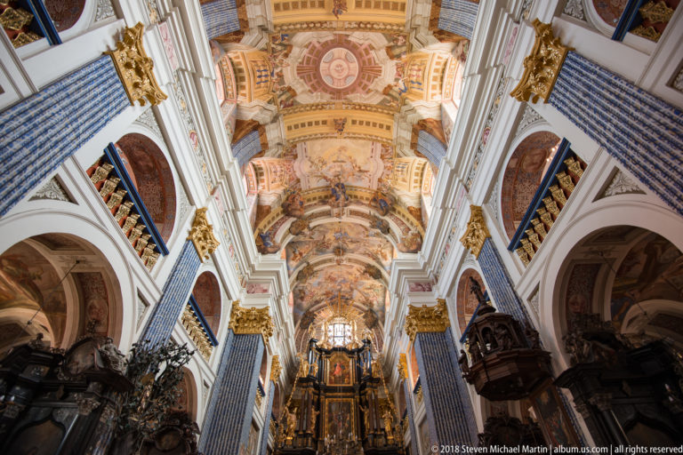 Long central nave view toward the high altar beneath frescoed vaults, with patterned pilasters, arches, dark furnishings, and chandeliers.