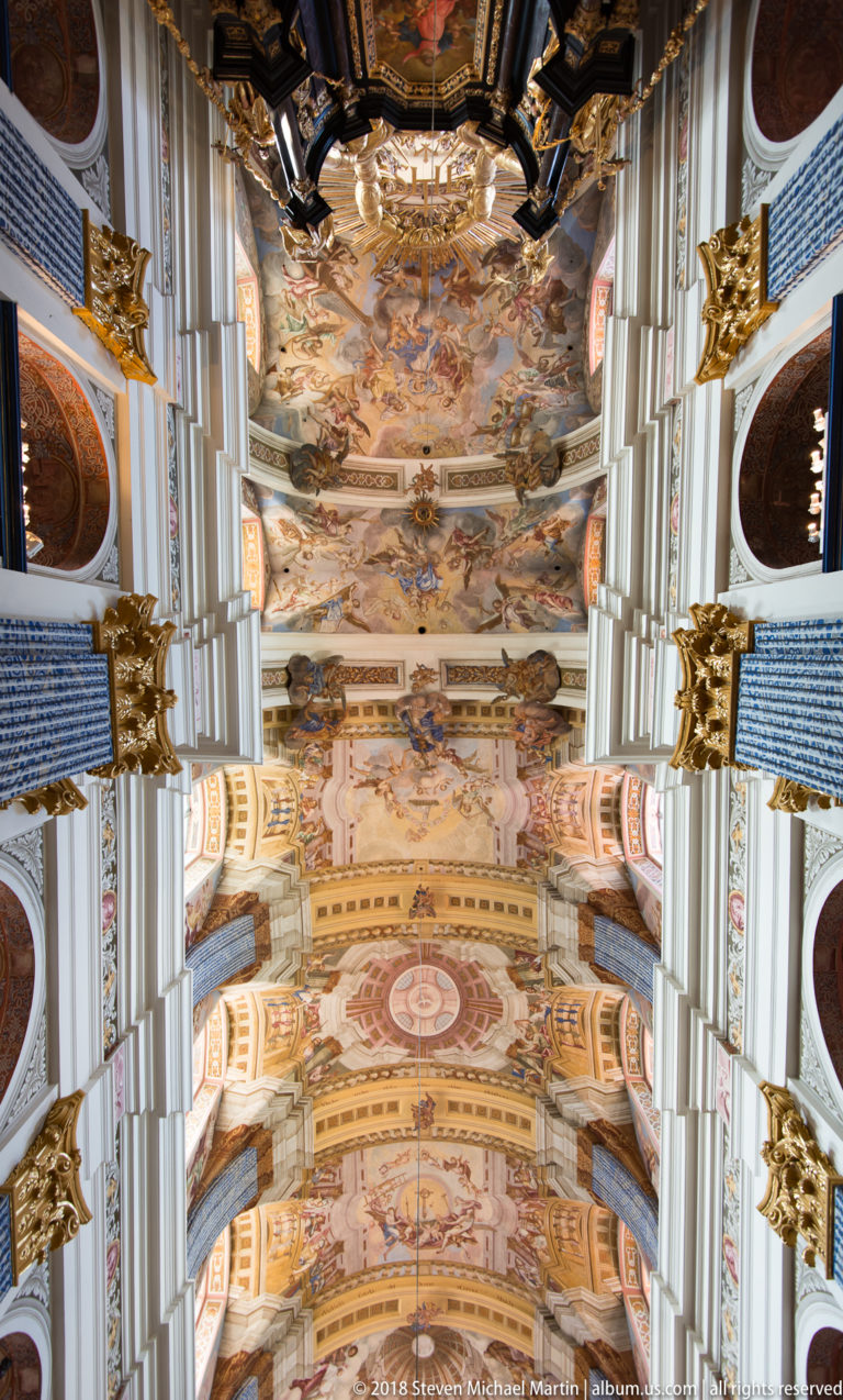 Centered upward view of successive frescoed vault bays framed by pale ribs, blue patterned pilasters, and gilded capitals.