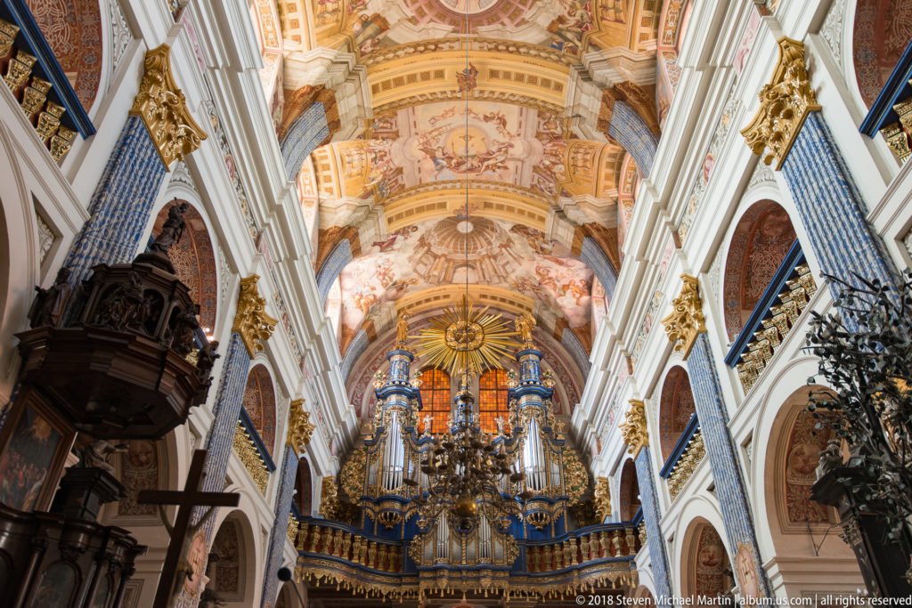 Large pipe organ beneath frescoed vaults, framed by tall white-and-blue nave architecture, with a dark pulpit and cross at left.