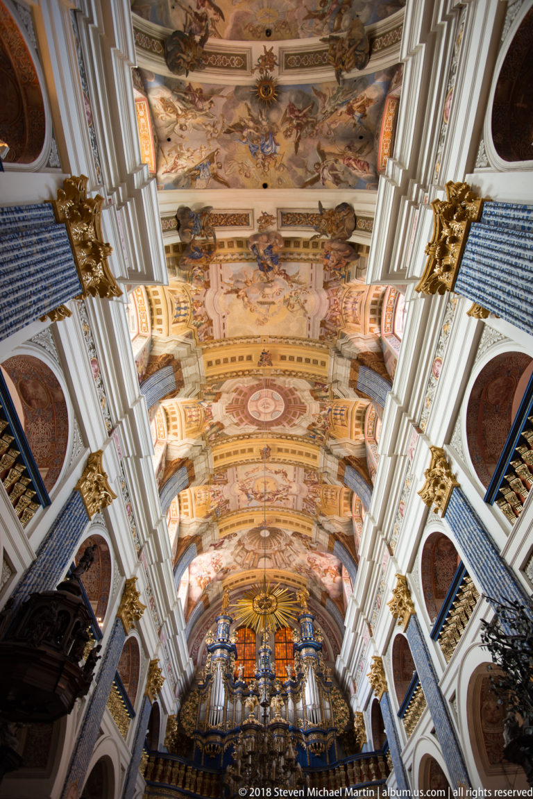 Upward nave view toward the pipe organ, with frescoed ceiling bays, white ribs, blue patterned pilasters, and gilded capitals.