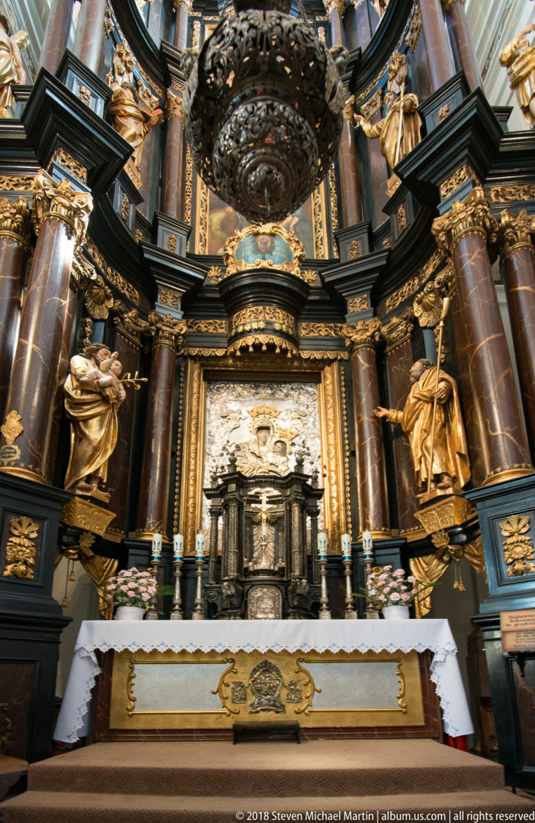 Close view of the high altar with tabernacle, suspended dark ornament, gilded figures, reddish columns, candles, and decorated frontal.