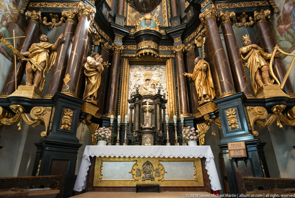 Silver-toned tabernacle centered between gilded standing figures and reddish columns, with candles, flowers, and a white altar cloth.