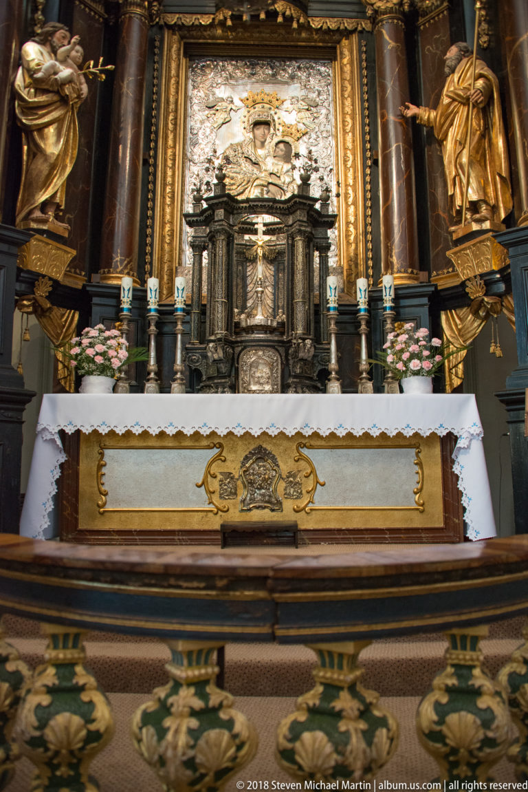 Frontal detail of the high altar with silver-toned tabernacle, relief panel, candles, white altar cloth, and dark columns.