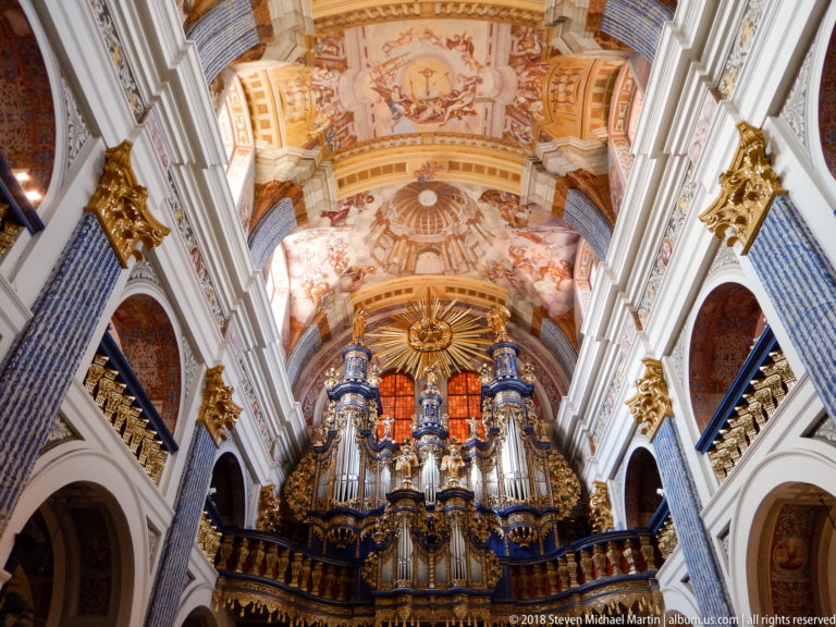 Axial view up the nave toward an ornate pipe organ framed by arcades, gilded capitals, and a frescoed vaulted ceiling.