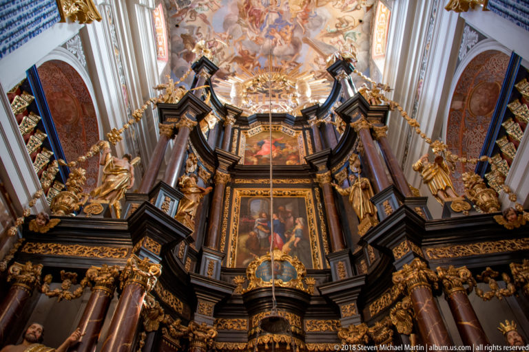 Tall dark-and-gold Baroque high altar with columns, framed sacred images, gilded figures, tabernacle, and frescoed vault above.