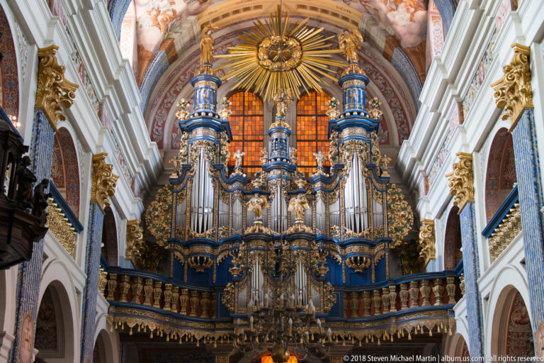 Frontal view of a large blue-and-gold Baroque organ with silver pipes, gilded sunburst, sculptural ornament, and glowing windows.