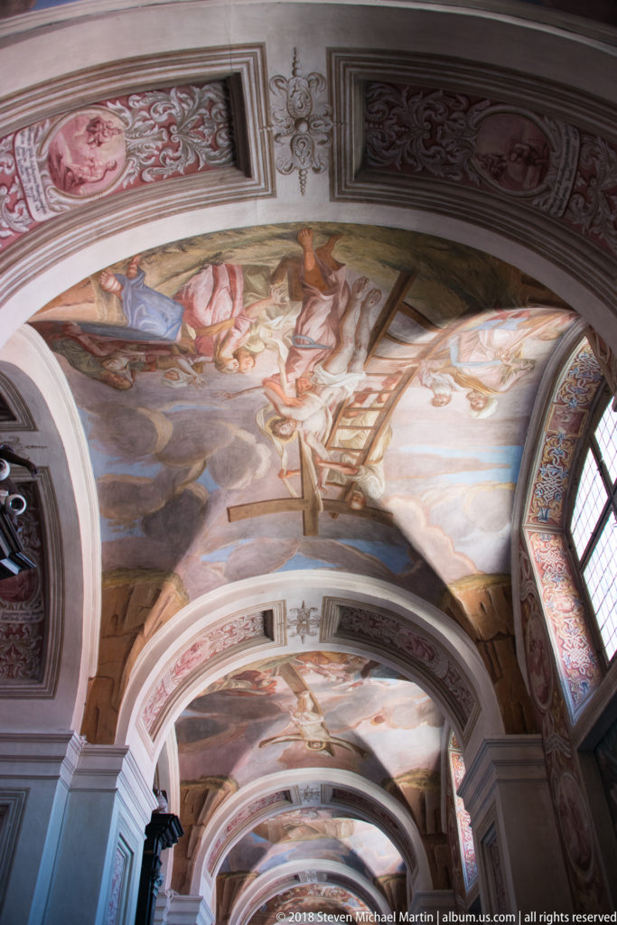 Oblique interior view across multiple frescoed vault bays divided by white arches, with a tall window at right.