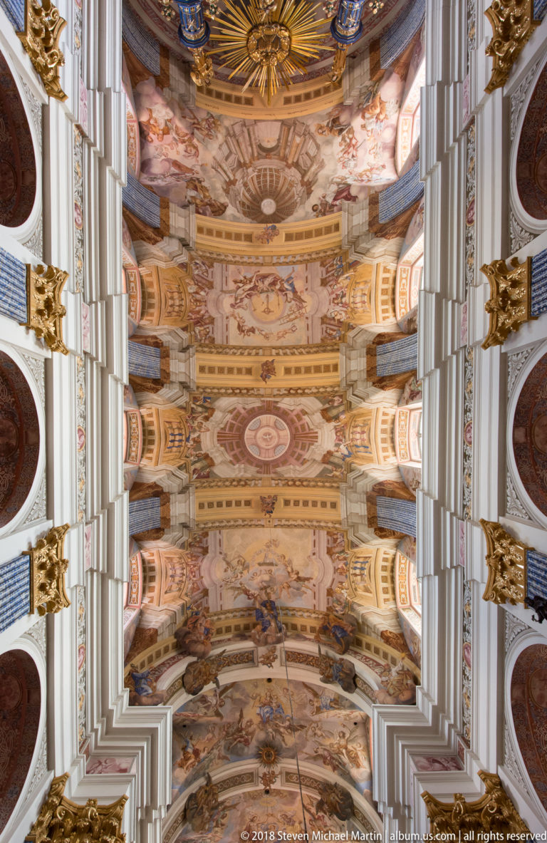 Straight-up symmetrical view of a frescoed nave ceiling divided by white ribs, with blue patterned supports and gilded capitals.