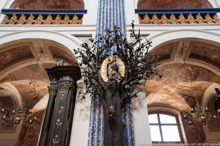 Dark branching metal devotional display with many small leaves and an oval sacred medallion mounted before a patterned pilaster.