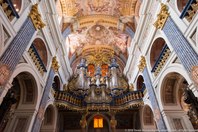 Baroque organ framed by white arches and blue patterned pilasters, with gilded details, ceiling frescoes, and an orange window.