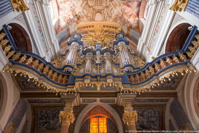 Close frontal view of a blue-and-gold Baroque organ with multiple pipe towers, gilded figures, sunburst, and gallery below.