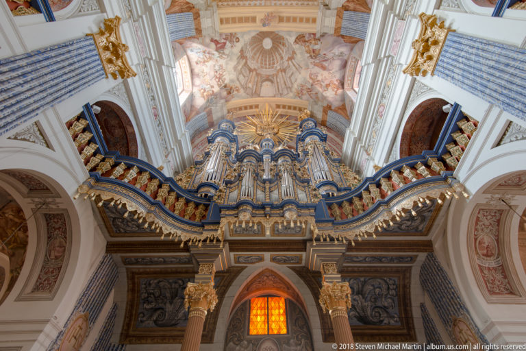 Large blue-and-gold pipe organ with silver pipes, gilded figures, gallery rail, sunburst, and frescoed ceiling above.