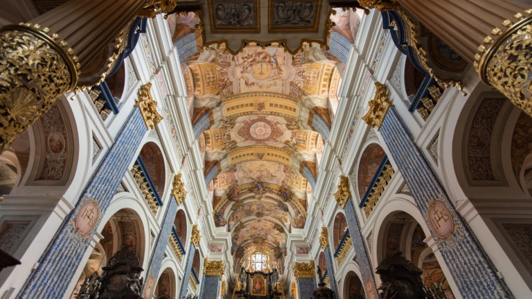 Upward nave view with frescoed vault bays, white ribs, blue patterned pilasters, gilded capitals, arches, and chandeliers.