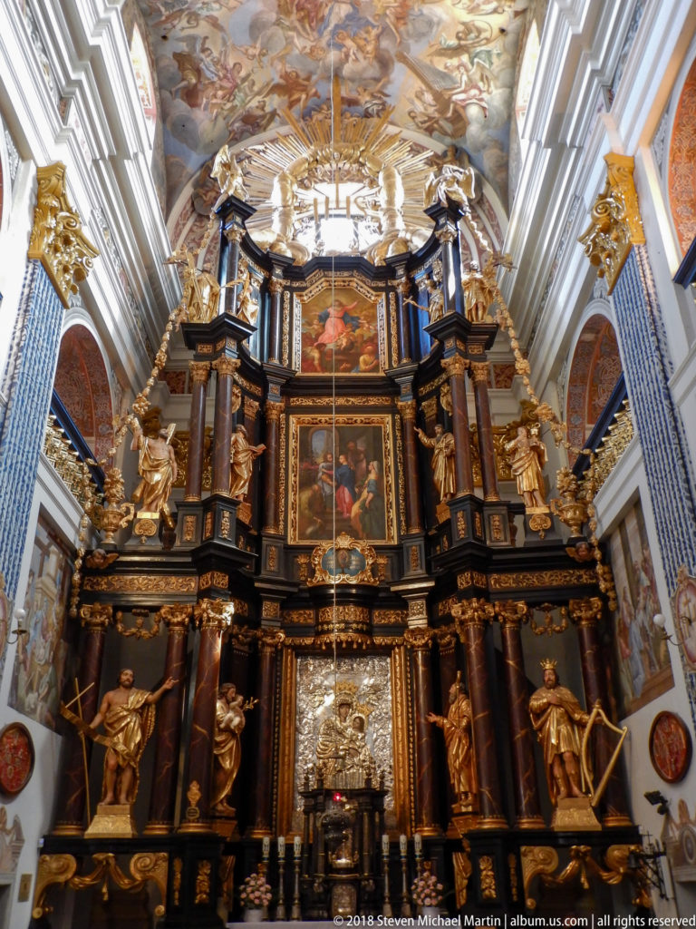 Monumental dark and gilded Baroque high altar with stacked religious paintings, sculptures, and a silver-toned centerpiece beneath frescoed vaulting.