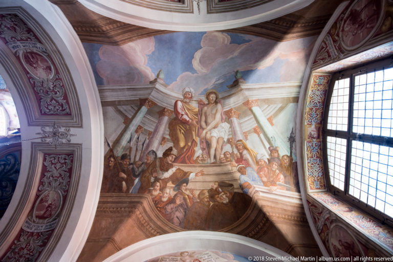 Ceiling fresco with two elevated central figures above a crowd, framed by curved moldings and a window at right.