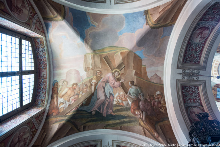 Baroque ceiling fresco with robed figures gathered around a large stone structure, beside a window and curved vault molding.