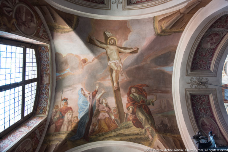 Crucifixion fresco with Christ on the cross and robed figures below, beside a tall window and painted vault framing.
