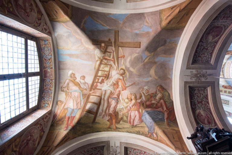 Ceiling fresco showing a tall cross and ladder above clustered robed figures, framed by vault ribs and a window.