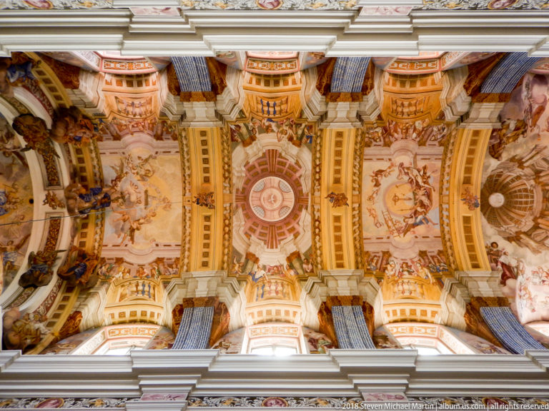 Upward view along an ornate Baroque nave ceiling with figurative frescoes, white and gold ribs, and illusionistic architectural decoration.