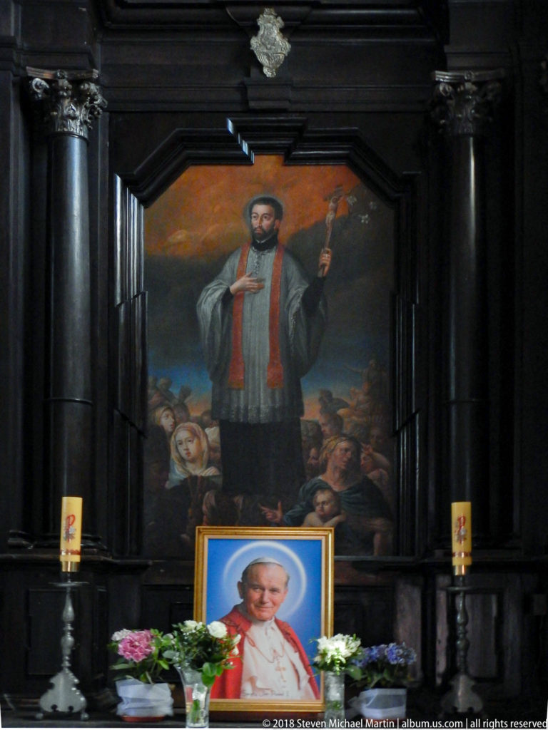 Dark side altar with a large painting of a robed saint above a framed portrait of Pope John Paul II, flowers, and candles.