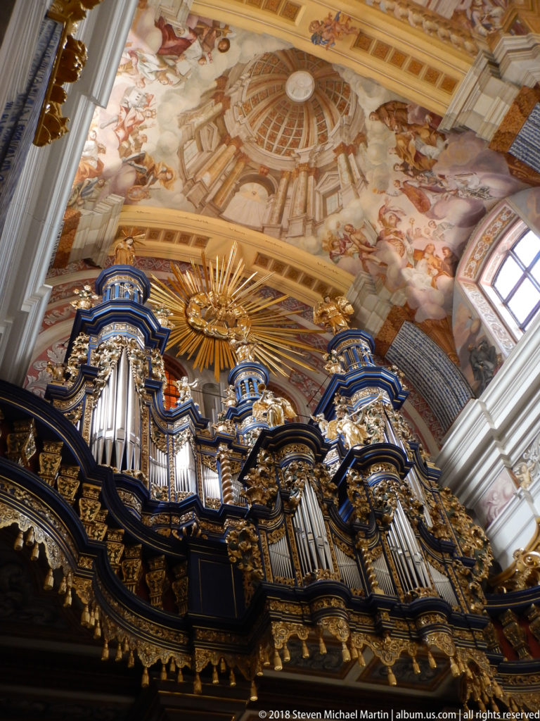 Large Baroque pipe organ with silver pipes, dark and gilded carving, blue accents, and a radiant sunburst beneath a frescoed ceiling.
