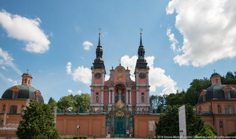 Distant frontal view of the pink twin-towered sanctuary behind its wall and gate, with corner pavilions, trees, and clouds.